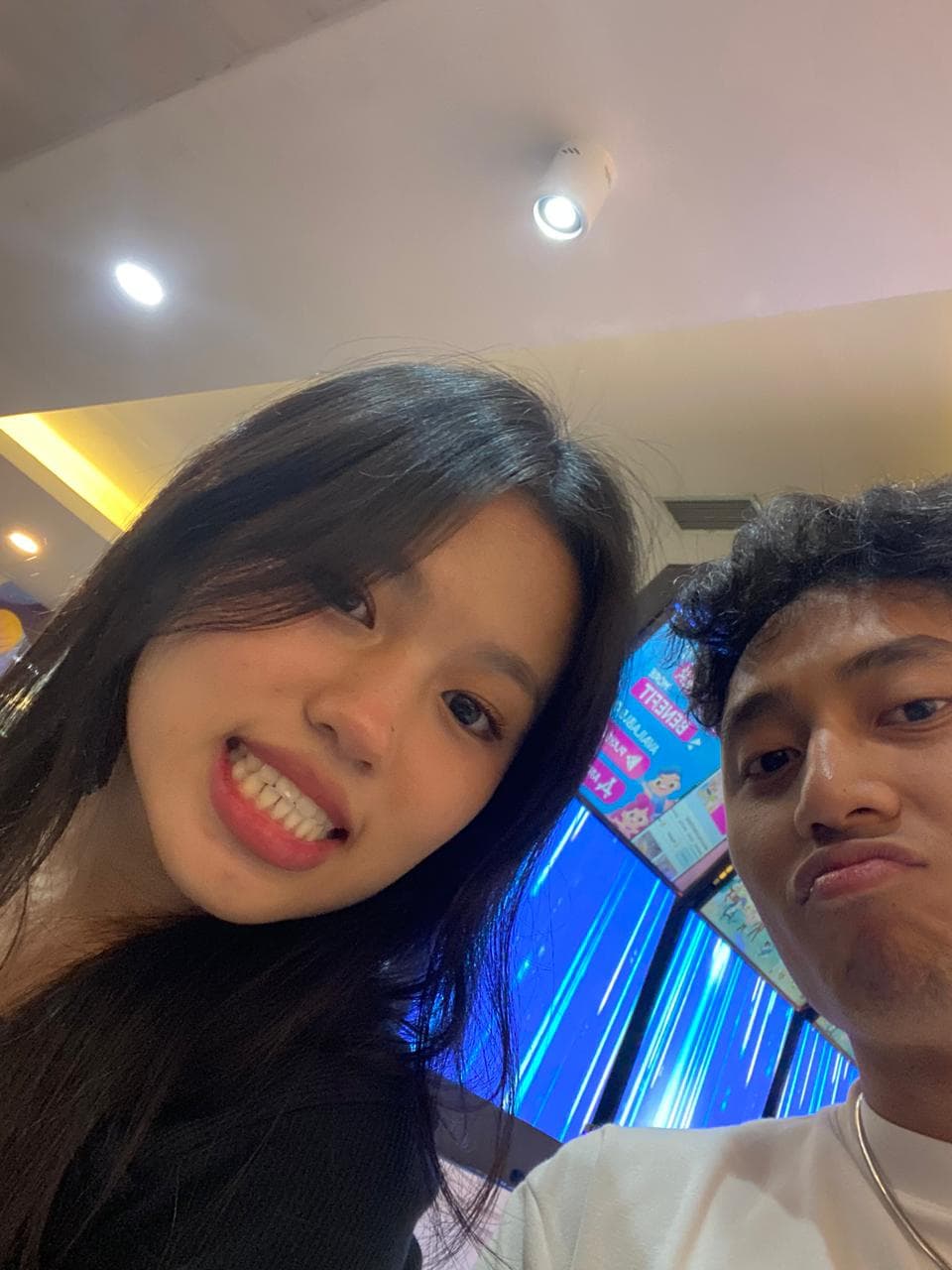 Nayla tersenyum dan pasangannya berpose playful dalam selfie di area arcade.