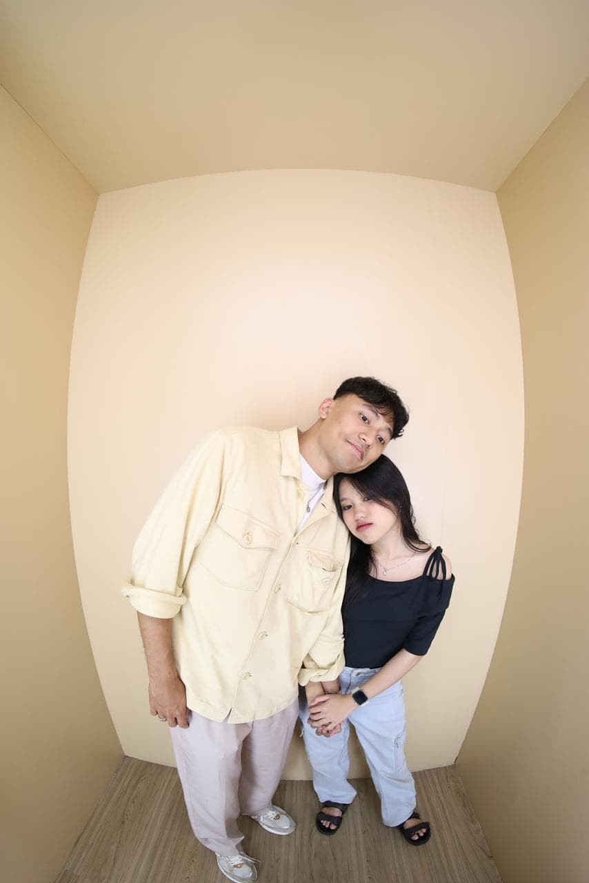 Pasangan berpose santai di ruang krem dengan kepala saling mendekat.