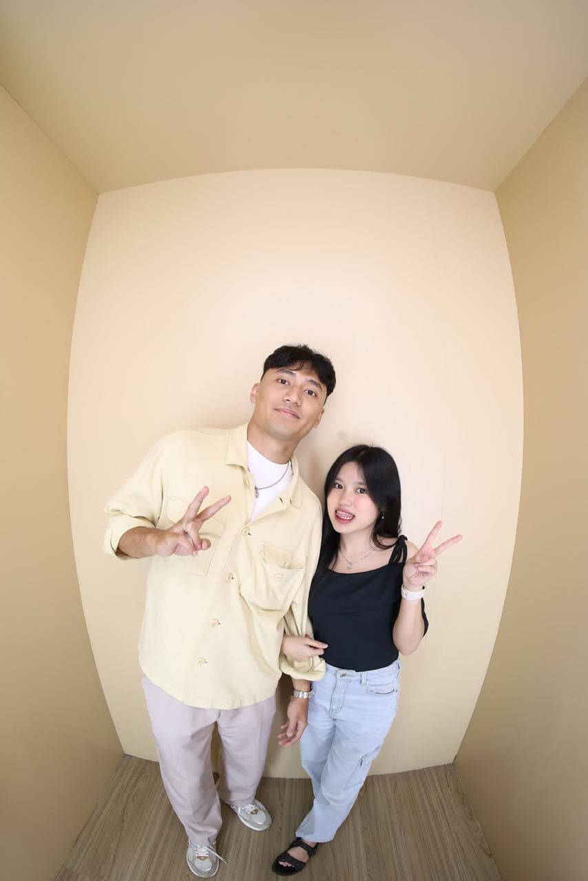 Nayla dan pasangannya membuat gestur peace sambil tersenyum di ruang krem.